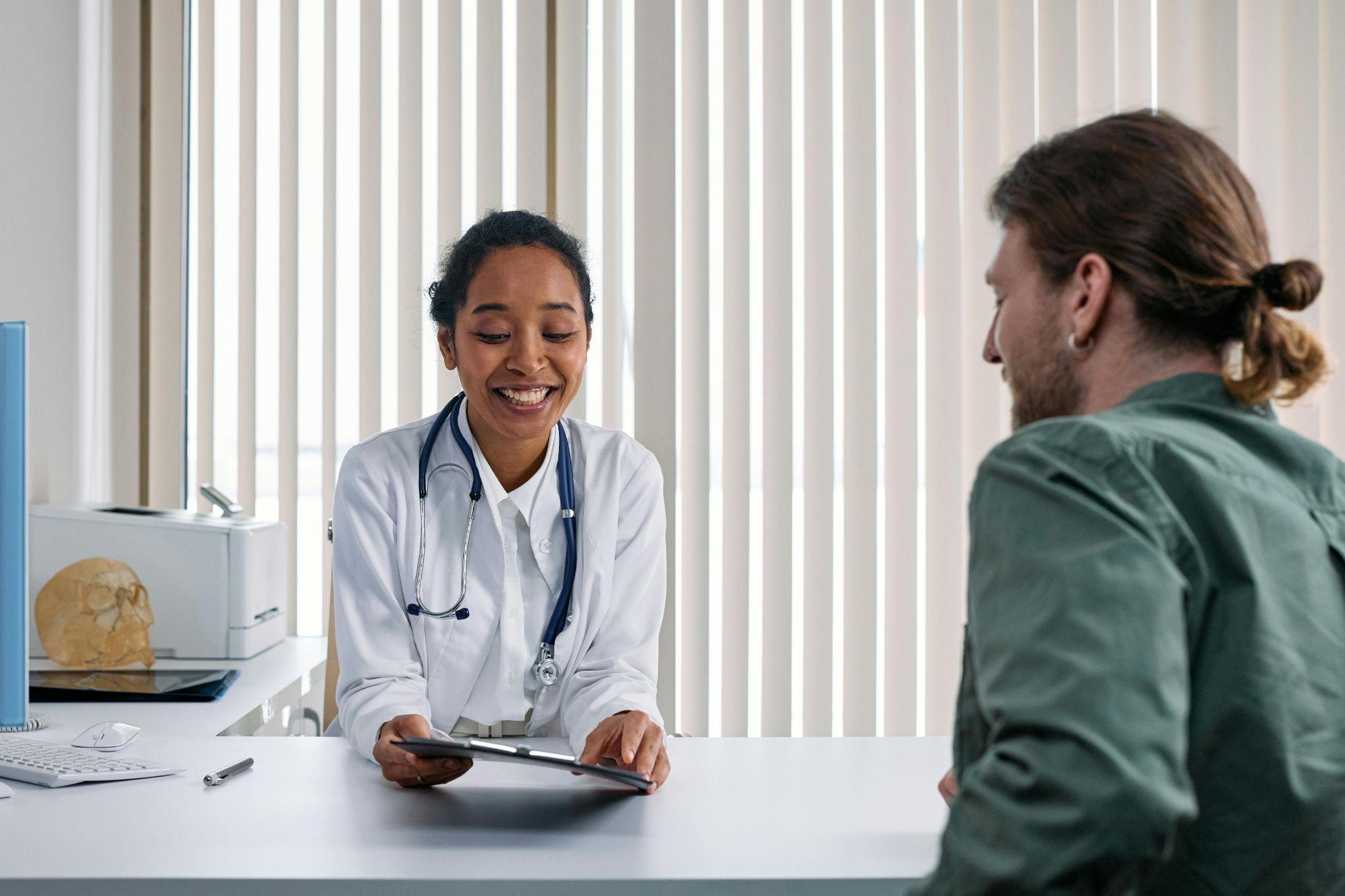 A smiling doctor sitting at a desk, holding a tablet and interacting with a patient