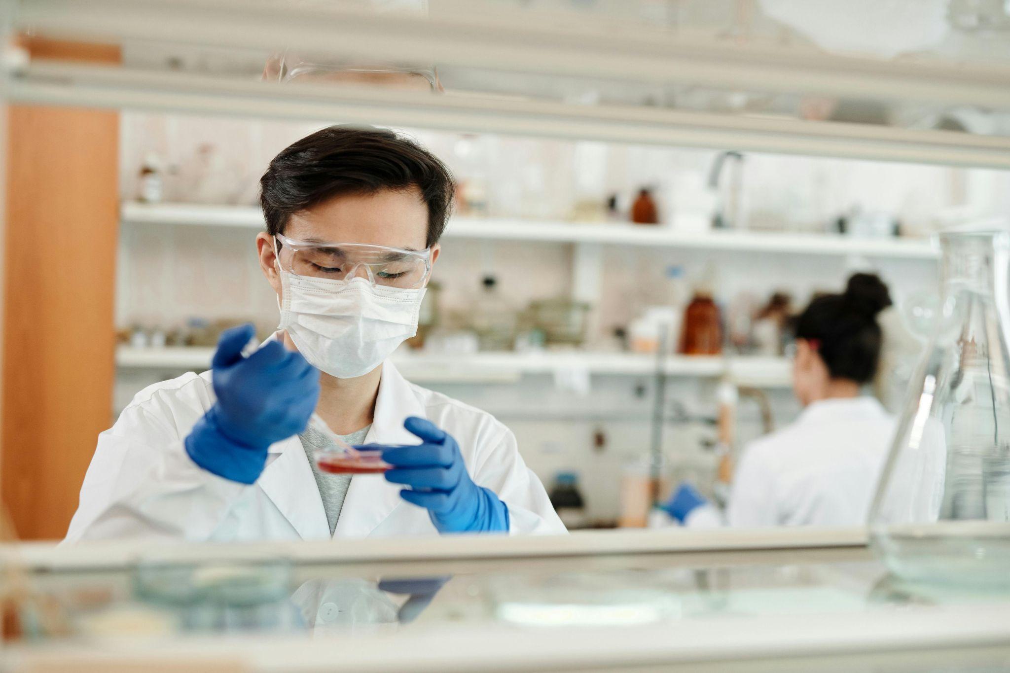 A lab technician wearing gloves and glasses, handling a petri dish, in a laboratory setting.