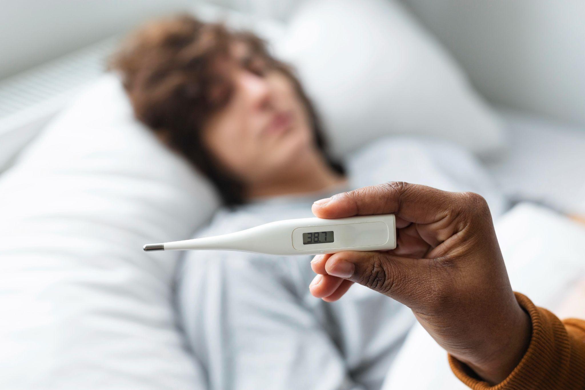 A person holding a thermometer showing a temperature of 38.7°C, indicating a fever.