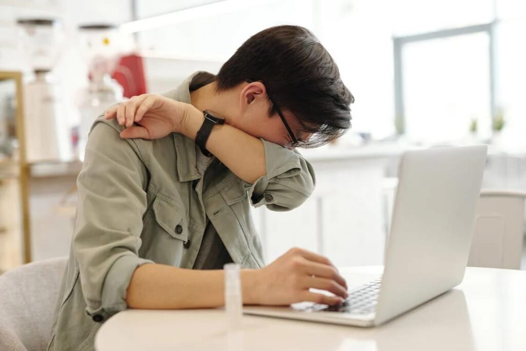 A man sneezing into a tissue while working on his laptop, indicating he needs a sick day