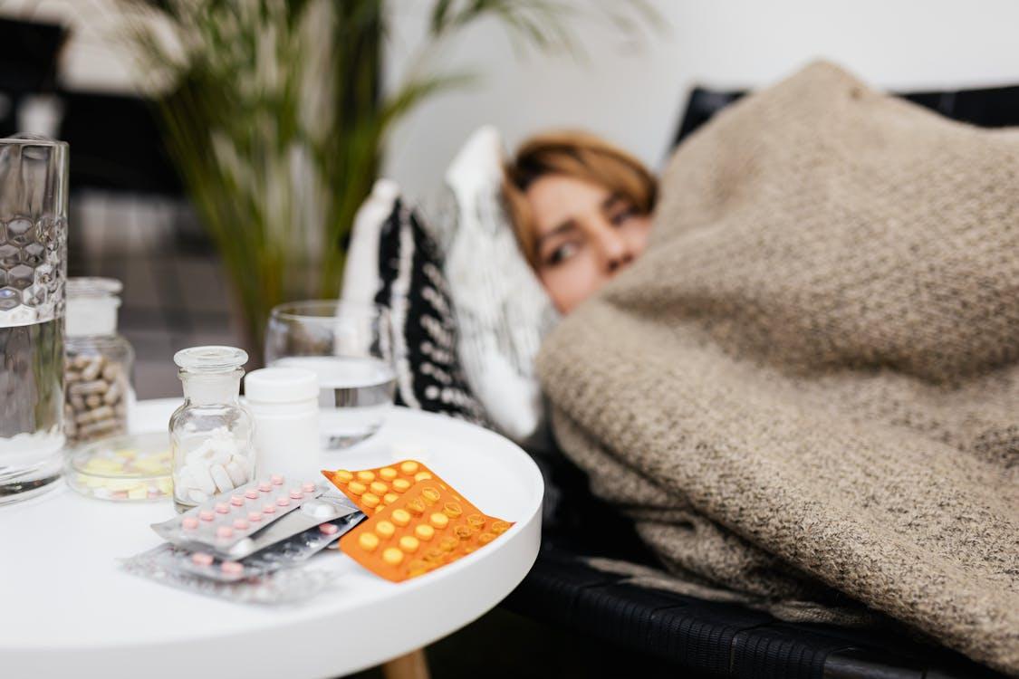 Various medicines on a table beside a woman resting under a blanket after taking sick leave