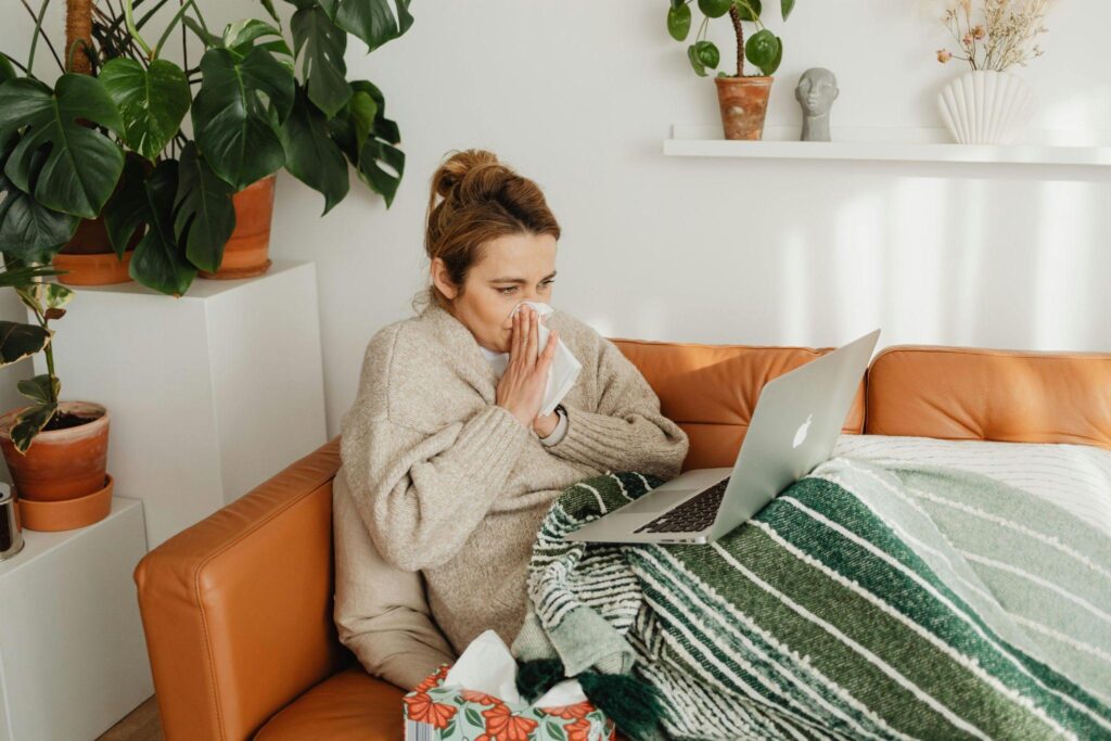 Woman sitting on couch under blanket using laptop while appearing unwell
