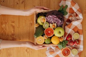 Hands holding a wooden box filled with assorted fresh fruits and vegetables.
