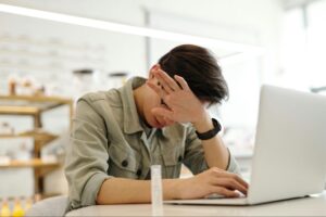 A man sitting at a desk, rubbing her forehead while working on a laptop.
