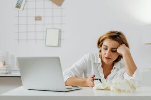 Woman working on a laptop at home while surrounded by tissues and appearing unwell.