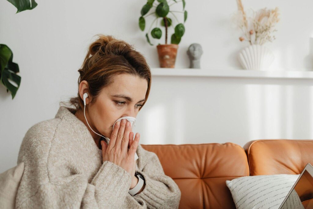 Woman sitting on a couch sneezing into a tissue while working on a laptop.