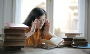 A woman sitting at a desk, rubbing her temples with books stacked around her.