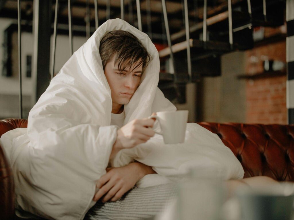 A tired person wrapped in a blanket, drinking a hot beverage to stay warm.