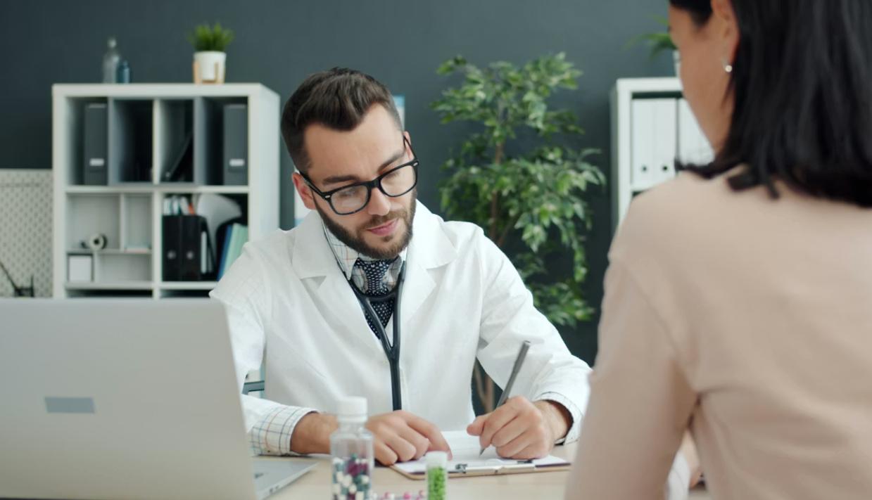 This image shows a doctor writing notes during a consultation at a desk.