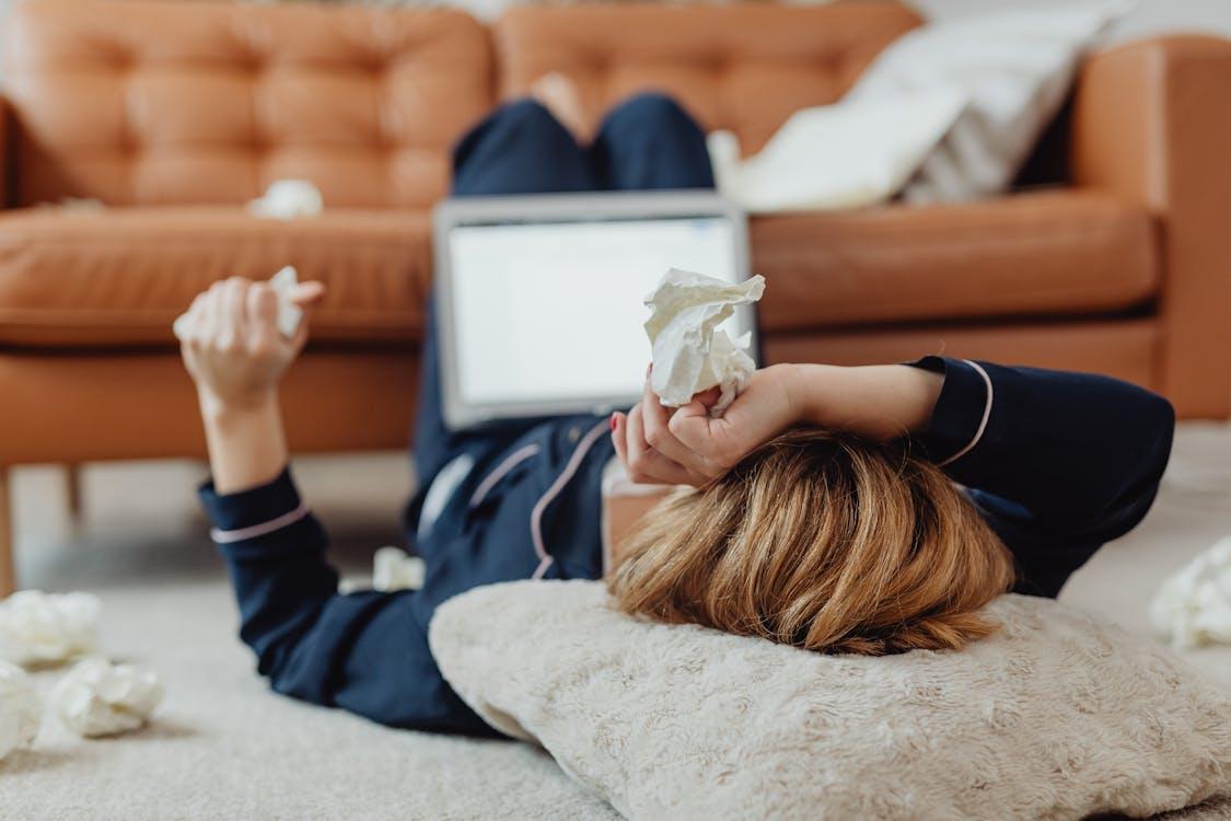 Adult lying on the floor with a laptop and tissues, showing exhaustion
