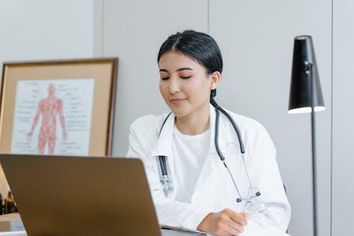 Doctor sitting in clinic writing a medical note on paper with a laptop nearby