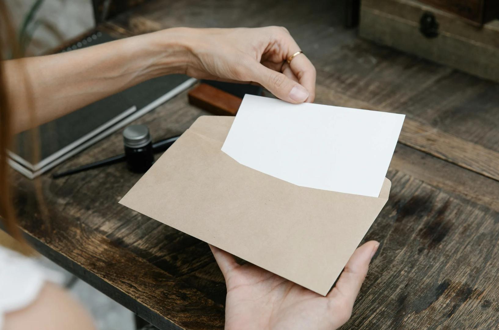 This photo shows hands holding a paper document and placing it into an envelope on a wooden desk.