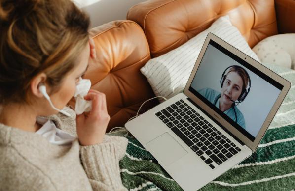 This image shows a person at home on a laptop speaking to a doctor via video call, holding tissue.